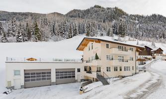A modern building in the snow, surrounded by a snowy landscape and tall fir trees. Mountains can be seen in the background.