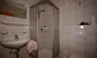 A small bathroom with a shower and a sink. The walls are tiled, and there is a shelf for toiletries.