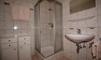 A small bathroom with a shower and a sink. The walls are tiled and there is a mirror as well as storage space.