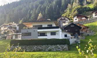 A modern house in the mountains, surrounded by a green meadow. In the background, other traditional buildings and forests can be seen.