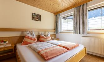 A cozy bedroom with a double bed and decorative pillows. There are large windows that provide plenty of natural light, as well as wooden flooring and a warm atmosphere.