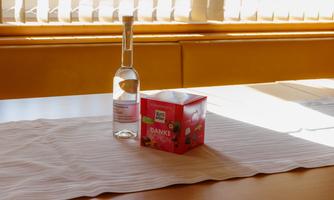A bottle of clear drink stands next to a colorful gift box on a table. The table is covered with a white cloth and light is coming through the windows.