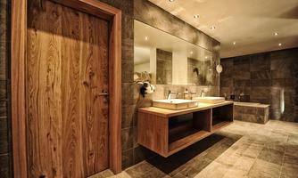 A modern designed bathroom with a wooden countertop and two sinks. The walls are clad with grey tiles, and there is an elegant wooden front door.