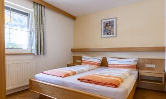 A cozy room with a double bed and bright, cushioned pillows. The room has a window with curtains and a decorative mural.