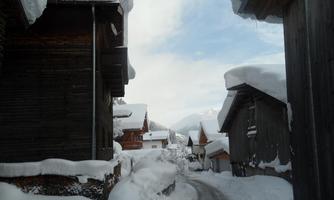 A snow-covered road between rustic wooden buildings. The sky is cloudy and the surroundings are idyllic and calm.