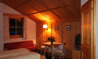 A cozy room with wooden paneling and warm light. There is a bed, a table, chairs, and a window view.