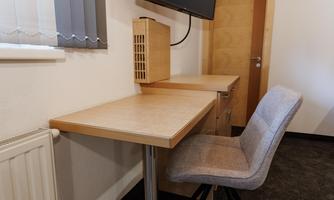A simple workspace with a wooden desk and a comfortable chair. In the background, a television is mounted on the wall.