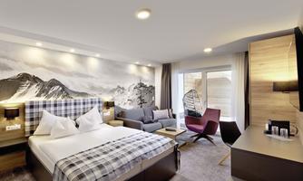 A modern hotel room with a large double bed and a cozy seating area. The walls feature a picturesque mountain landscape, and the room has access to a balcony.