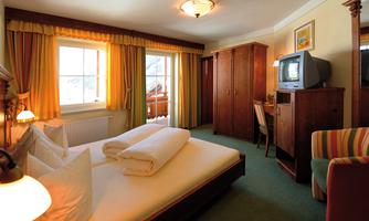 A cozy bedroom with a double bed, a television, and a desk. Large windows with curtains provide plenty of light.