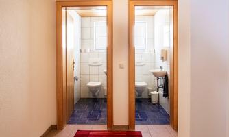 A hallway with two open bathroom doors. The bathrooms are bright and modernly furnished, with white tiles and toilets.