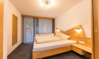 A modern bedroom with a large bed and wooden shelves. The large windows are equipped with blinds and provide various lighting conditions.