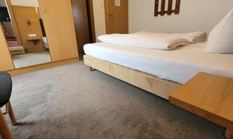 A simple bedroom with a bed, a table, and a mirror. The walls are light, and there is a wooden clothes rack.