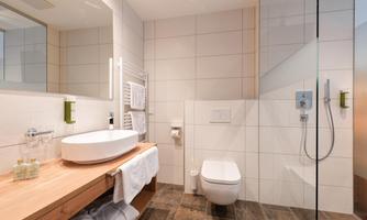 A modern bathroom with a light tiled floor and a glass shower cubicle. On the wooden shelf are care products and fresh towels.