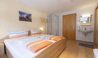 A modern room with two beds, bright walls, and a warm wooden floor. In the background, a shower and sink are visible, as well as a decorative picture on the wall.