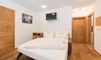 A modern room with a double bed, wooden floor, and a television on the wall. The walls are painted in light colors, creating a friendly atmosphere.