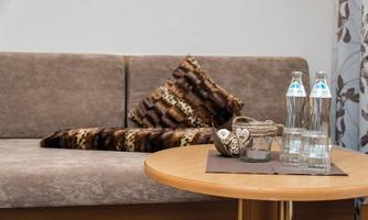 A cozy living room with a beige sofa and decorative cushions. On a wooden table, there are two glass bottles and a decorative container.