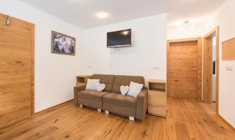 A bright living space with a comfortable sofa and a television on the wall. The floor is made of wood and the walls are painted in neutral colors.