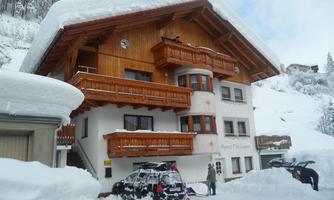 A chalet-style building in the snow with wooden balconies. The surroundings are characterized by snow-covered trees.