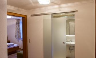 A bright hallway with a closed glass cabinet and access to a bathroom. In the background, a bedroom with a window and hardwood floor can be seen.