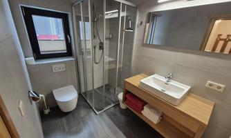 A modern bathroom with a shower and sink. The walls are tiled light gray, and there is a wooden cabinet with towels.