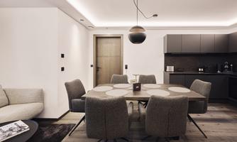 A modern dining room with a large table and chair-like seating. The walls are in neutral colors, and there is an elegant kitchenette in the background.