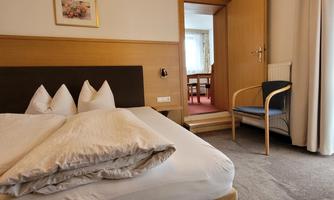 A cozy bedroom with a large bed and white pillows. In the background, there is a chair and a door leading to another room.
