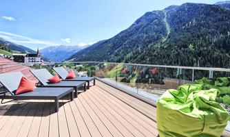A cozy terrace with loungers and colorful cushions. In the background, majestic mountains stretch under a clear blue sky.