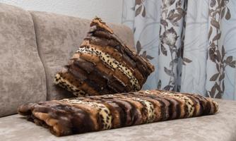 A cozy sofa with a decorative cushion and a soft blanket in brown tones. The background features a curtain with a floral pattern.