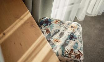 A colorful stool with floral patterns stands next to a wooden shelf. The room is brightly lit by the window.