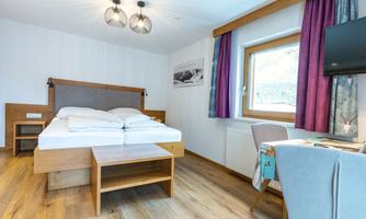 A modern bedroom with a double bed and a small table. The windows are decorated with curtains and there is a television on the wall.