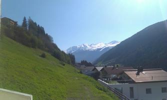 A picturesque mountain landscape with snow-covered peaks and green meadows. In the foreground, some houses are visible.