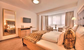 A modern bedroom with a large bed and a mirror. The windows let in a lot of light and there is comfortable seating.