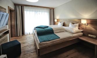 A modern bedroom with wooden decor and large windows. It has a double bed, a cozy seating area, and plenty of daylight.