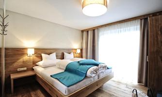 A modern bedroom with a cozy double bed and bright wood details. Large windows provide plenty of daylight and an inviting atmosphere.