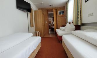 A cozy room with two single beds and a television mounted on the wall. The room has a bright atmosphere and leads to another room.