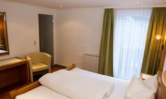 A cozy hotel room with a double bed, a table, and a chair. Large windows with curtains provide daylight and a welcoming atmosphere.