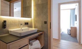 A modern bathroom with a white sink and wooden shelves. In the background, a bright hallway with a room is visible.