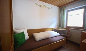 A simple bedroom with a single bed and a table. The room has a light-flooded atmosphere and a wooden floor.