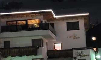 A charming building with a lit balcony in a wintry setting. There are parked cars and it is night.