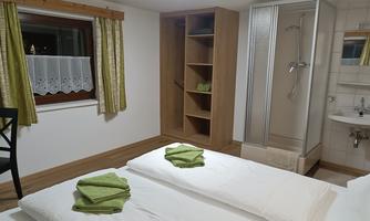 A simple bedroom with two beds, fresh towels, and a bathroom with a shower. The walls are painted in neutral colors, and there are green curtains.