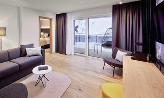 A modern living room with a gray sofa, a small table, and large windows. The room features a light wooden floor and a stylish decor.