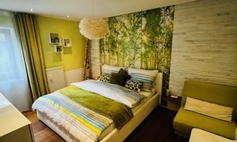 A modern bedroom with a large bed and colorful cushions. The walls are decorated with a calming forest motif, and there is a small sitting area.
