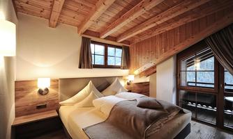A cozy bedroom with wood-paneled walls and a visible roof. The room is furnished with a double bed and large windows that provide plenty of daylight.