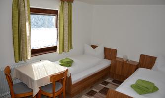 A simple room with two beds, a table, and chairs. The room has bright curtains and a view of the outside.