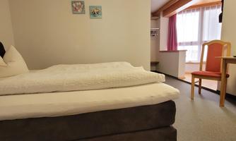 A cozy bedroom with a large bed and a chair. The room has light walls and a window with curtains.