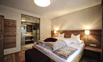A modern bedroom with a large bed and wooden details. In the background, a stylish bathroom is visible.