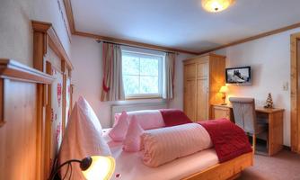 A cozy bedroom with two beds and wooden paneling. The room has windows overlooking the outdoors and a small desk.