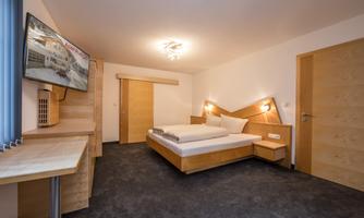 A modern bedroom with a double bed and a wooden wardrobe. The room is bright and equipped with a television and a desk.