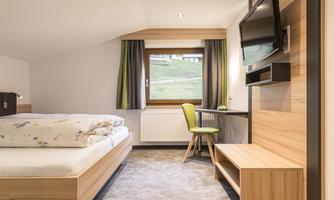 A modern hotel room with a double bed and a table. There is a television and a view of nature through the window.