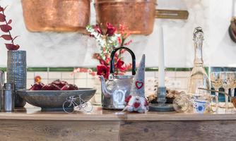 An inviting kitchen table with various kitchen utensils, a silver teapot, and fresh apples in a bowl. In the background, copper pots and a stylish vase are hanging.